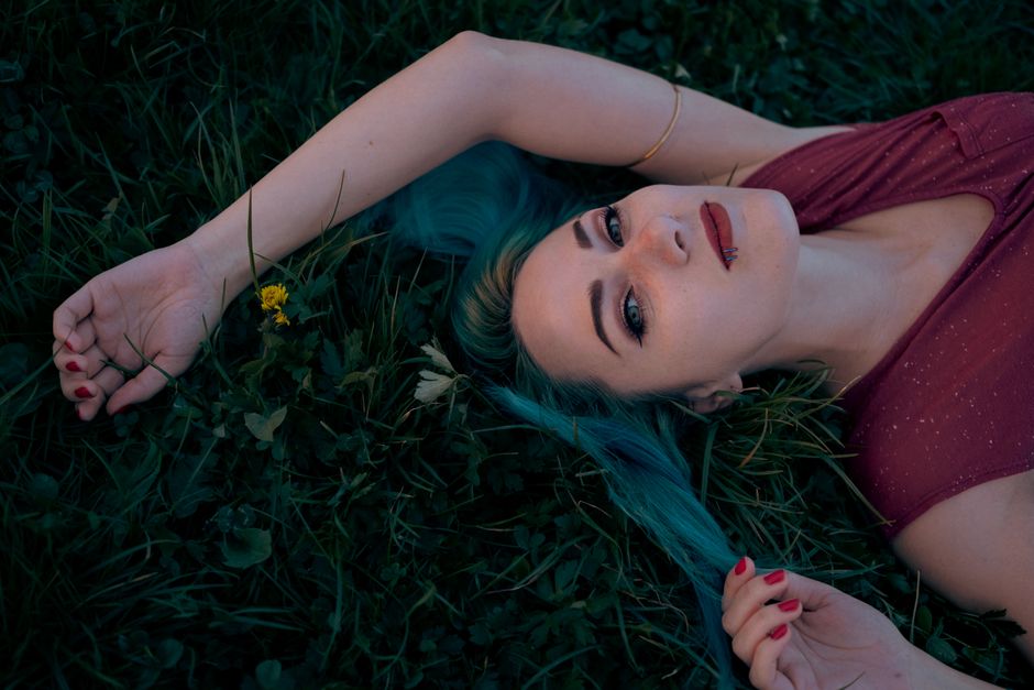 Woman with blue hair lying on grass in a rural meadow, expressing relaxed mood.