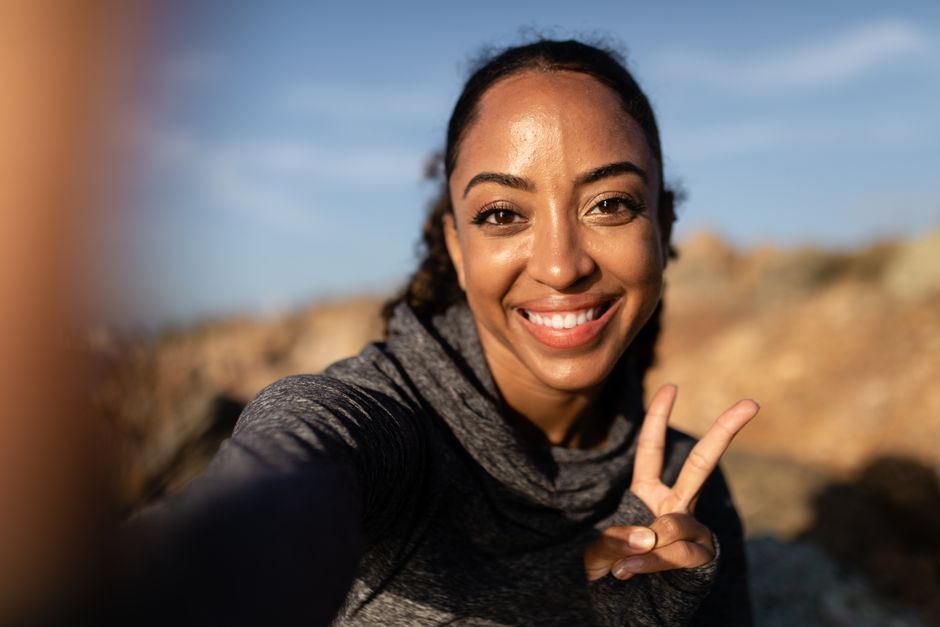 Smiling woman in grey hoodie taking a selfie outdoors, making a peace sign.