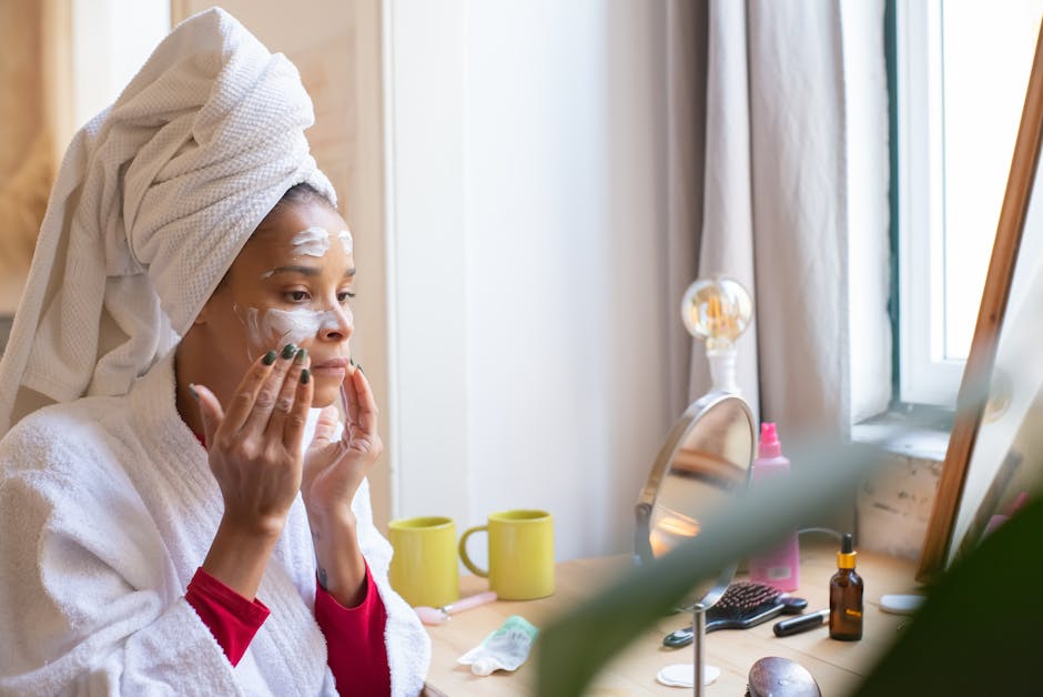 A woman applies face cream in a relaxing home setting, dressed in a bathrobe and head towel.