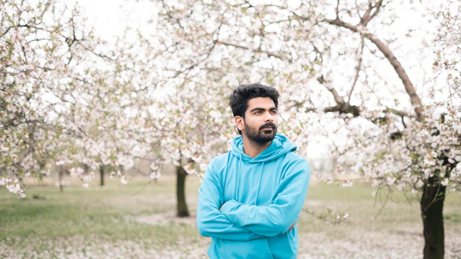 A man wearing a blue hoodie stands with crossed arms in a blooming orchard, exuding a serene springtime vibe.