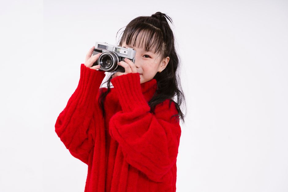 A young girl in a red sweater taking a photograph with a vintage camera.