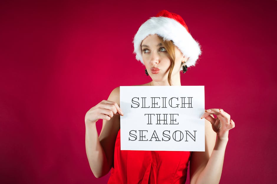 Young woman in Santa hat holding a 'Sleigh the Season' sign against red background.