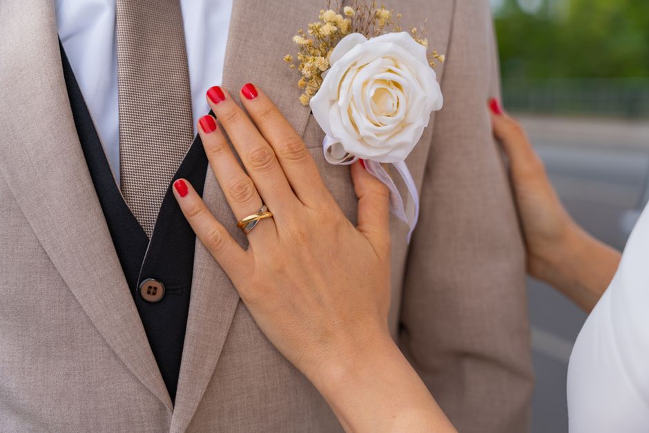 Close-up of a groom's suit with floral boutonniere and bride's hand showing wedding ring.