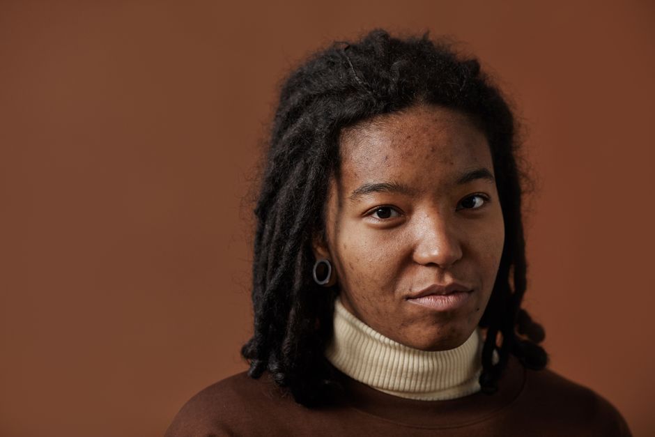 Close-up portrait of a young woman with dreadlocks, neutral expression, against a brown background.