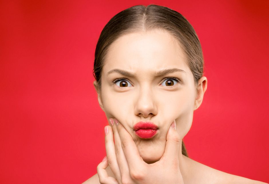 A young woman squeezes her cheeks, making a funny face against a vivid red background.