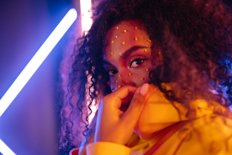 Artistic portrait of a young woman with colorful makeup and curly hair, captured in vibrant lighting.