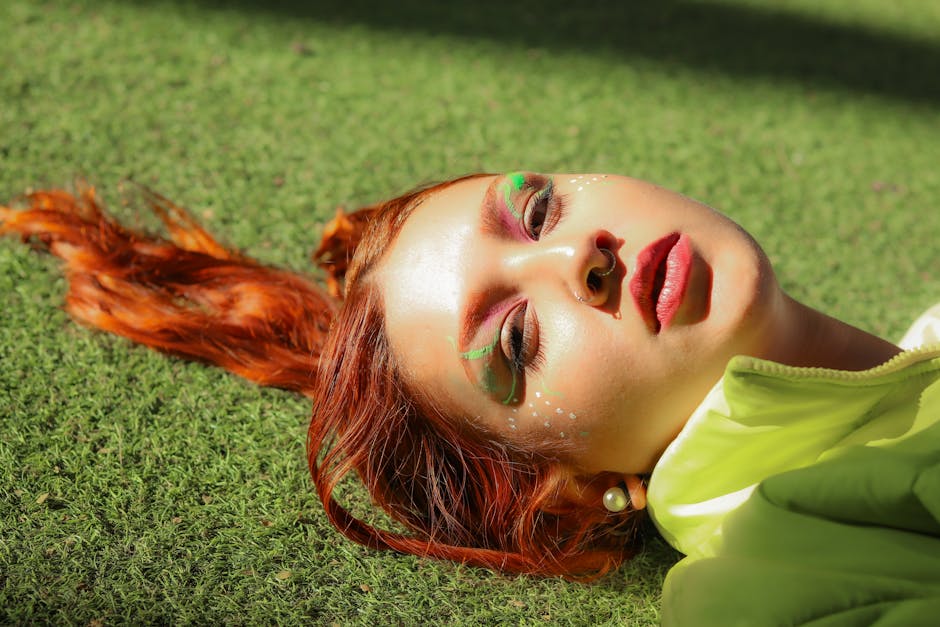 Vibrant portrait of a woman lying on grass with colorful eye makeup and red hair.