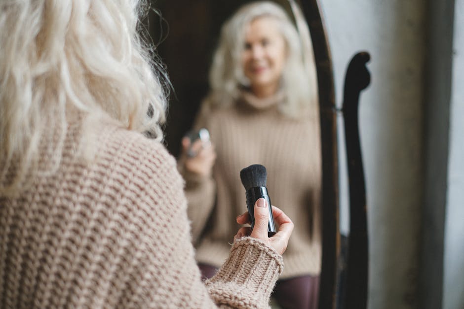 A senior woman applies makeup while looking in a mirror, holding a brush in her hand.