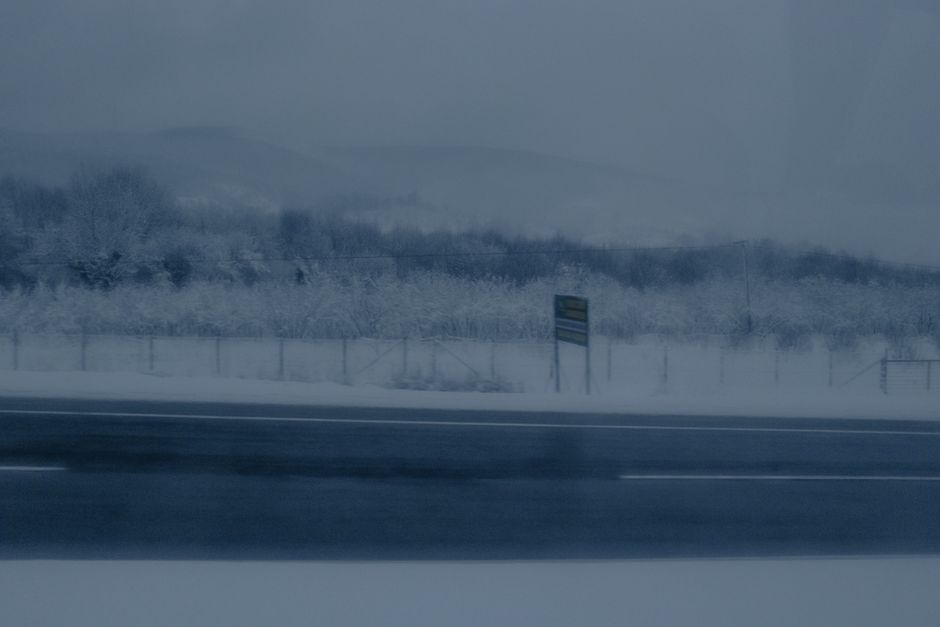 A moody winter scene showing a road, fog, and snow-covered trees during the day.