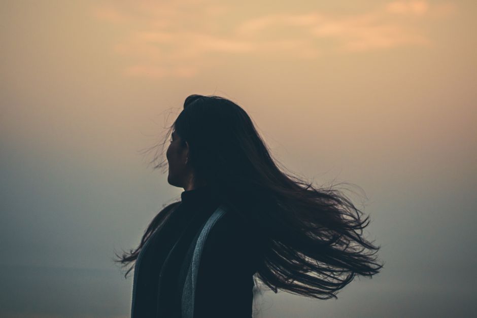 Side view of unrecognizable female shaking long hair while standing on shore near foggy sea on evening time in nature