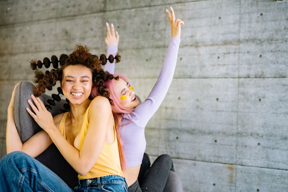 Two women with creative hairstyles and makeup pose happily against a concrete wall indoors.