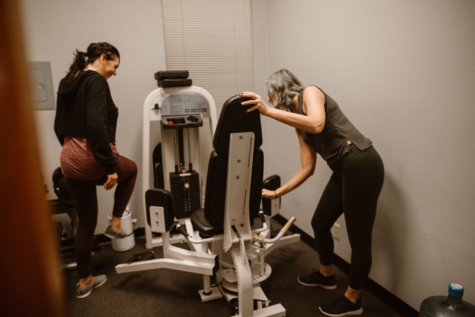 Two women exercising in a gym, adjusting and using a weight machine for fitness training.