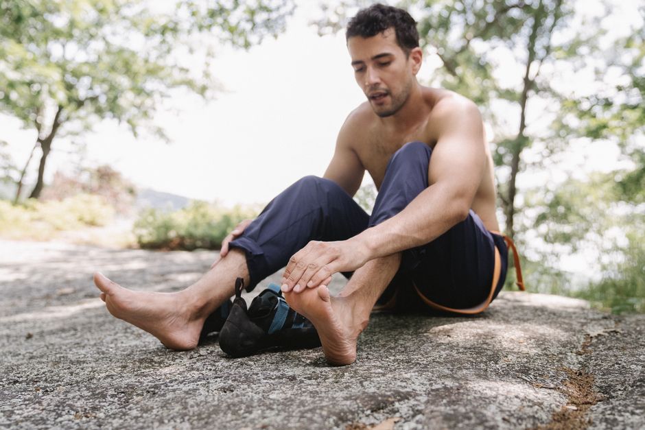 Shirtless man sitting on rock adjusting climbing shoes in sunny outdoor scene.