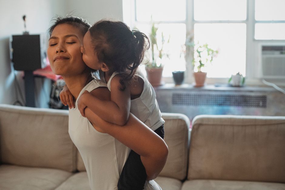 Side view positive young Asian woman piggybacking adorable daughter and getting cheek kiss while having fun together in light living room