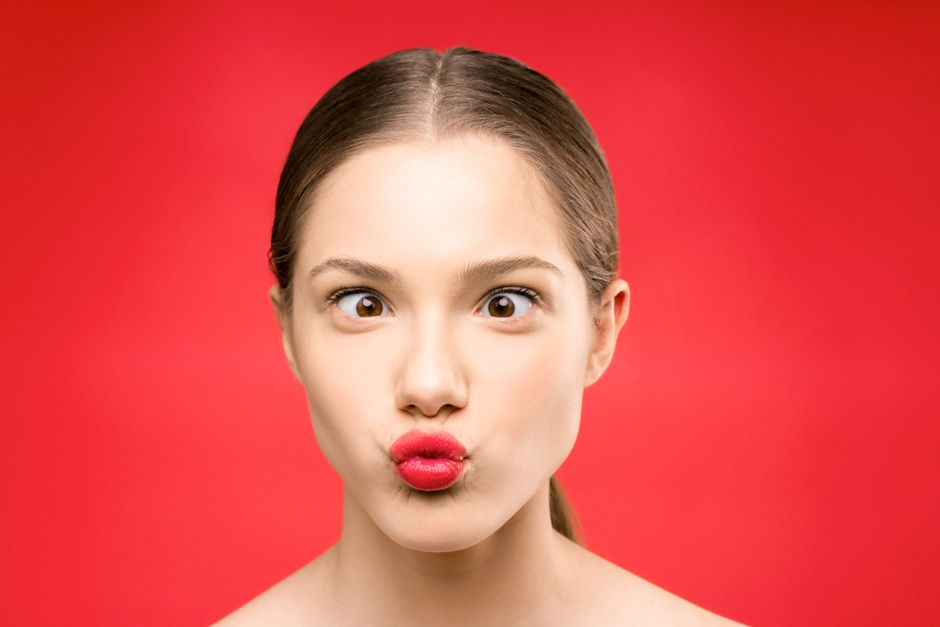 Young woman making a playful pout with bright red lipstick against a vibrant red background.
