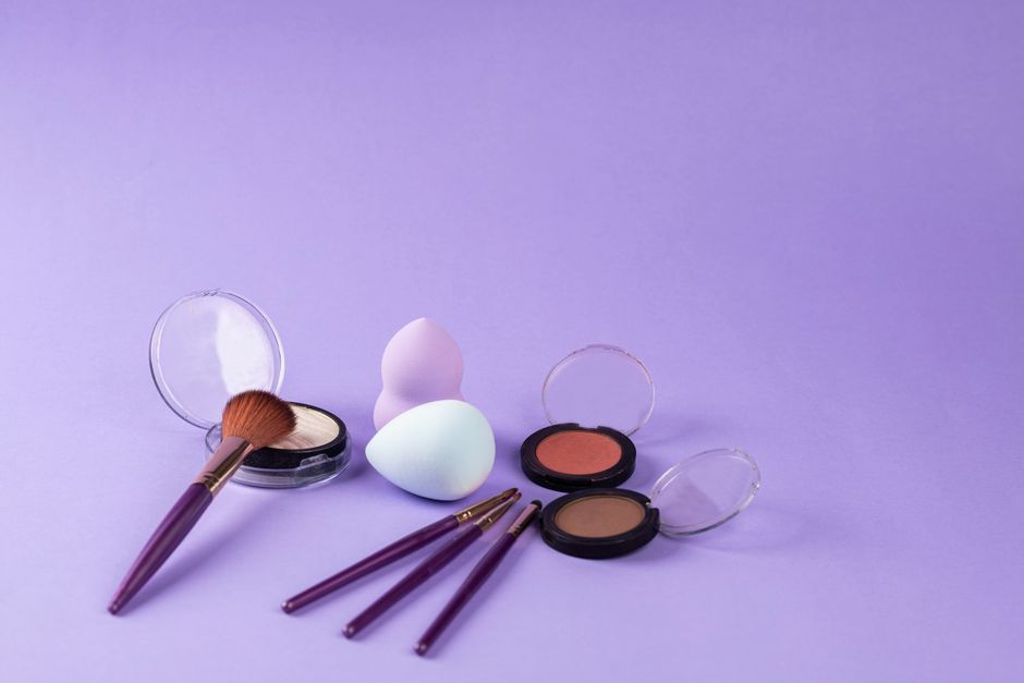 A colorful arrangement of makeup products including brushes and beauty sponges on a purple background.