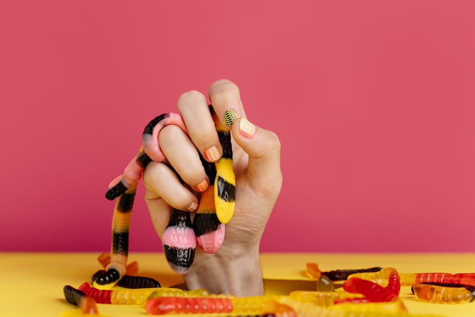 Close-up of a hand with manicured nails gripping colorful gummy worms against a pink background.