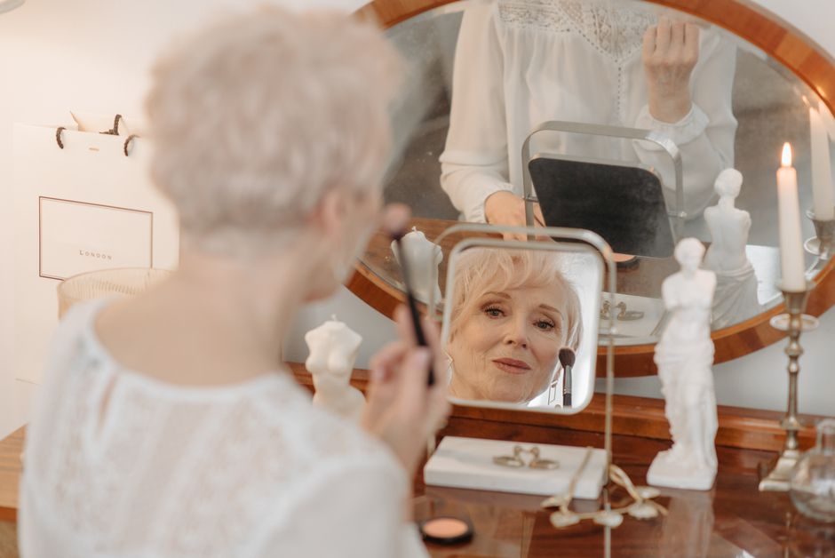 A senior woman applies makeup while looking in a vintage vanity mirror, creating a reflective and serene atmosphere.
