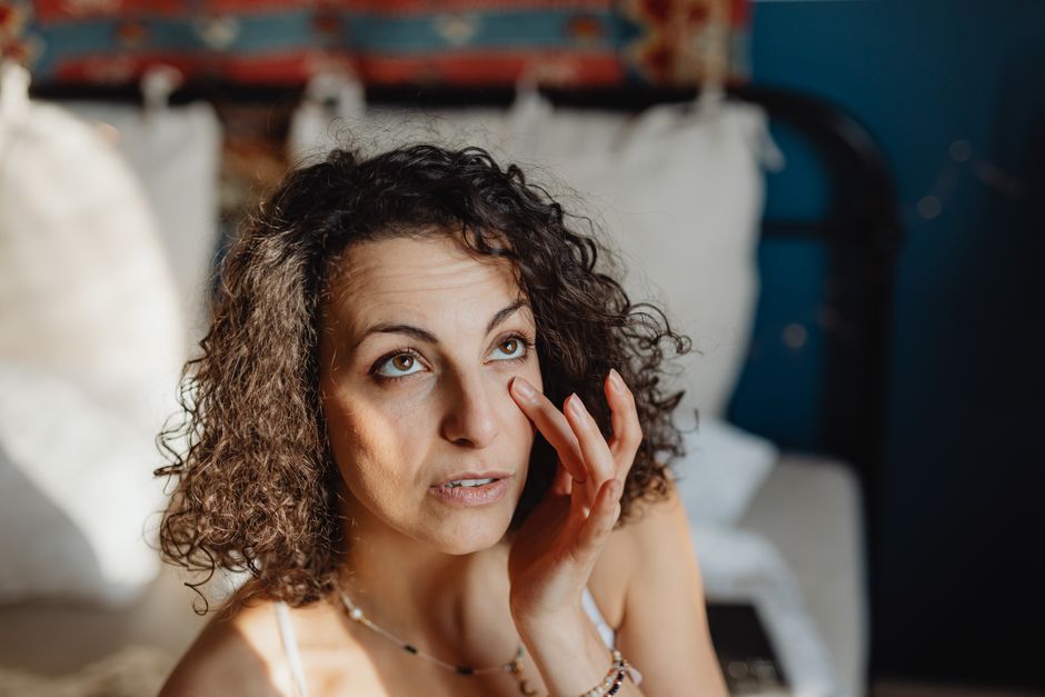 Woman applying skincare in a sunlit bedroom, embracing morning relaxation and self-care.