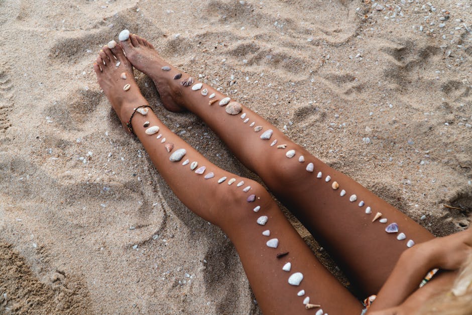 From above of skinny tanned legs of crop unrecognizable barefoot female covered with white pebbles sitting on sandy beach