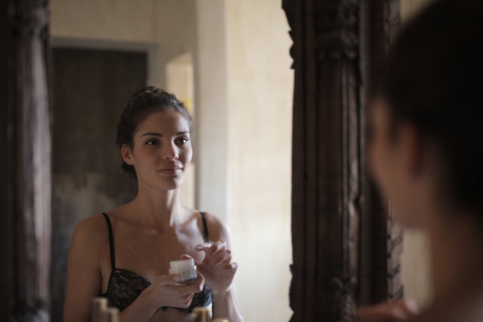A woman in lingerie applies cream while looking at her reflection in a mirror.