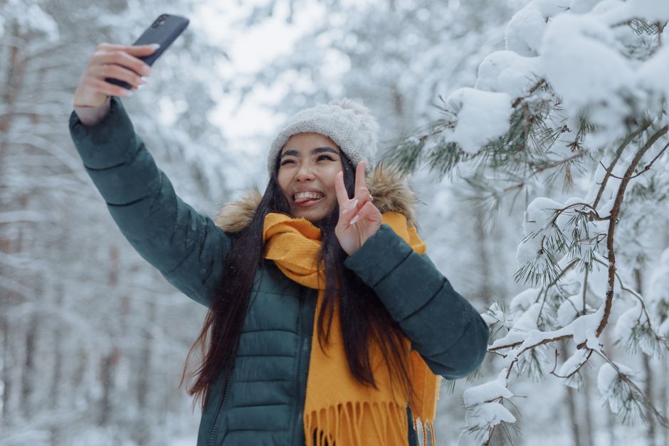 Smiling woman taking a selfie in a snowy winter forest, wearing a warm jacket and scarf.